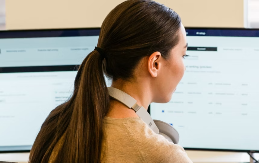 Woman with a ponytail and headphones around her neck looking at two monitors of a software interface