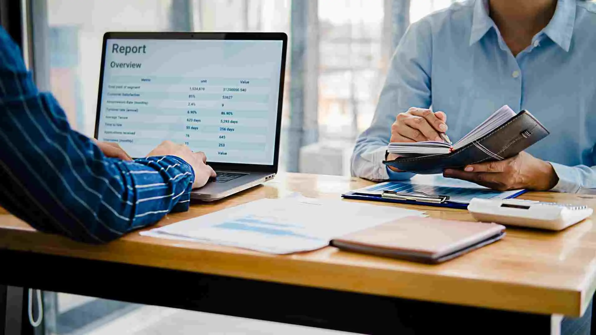Two people at a desk, one typing on a laptop showing a Report Overview table, the other taking notes