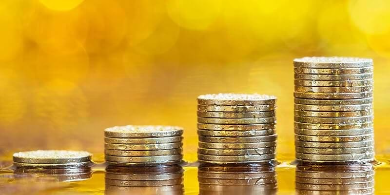 Four stacks of gold coins of increasing height on a reflective surface with a golden bokeh background