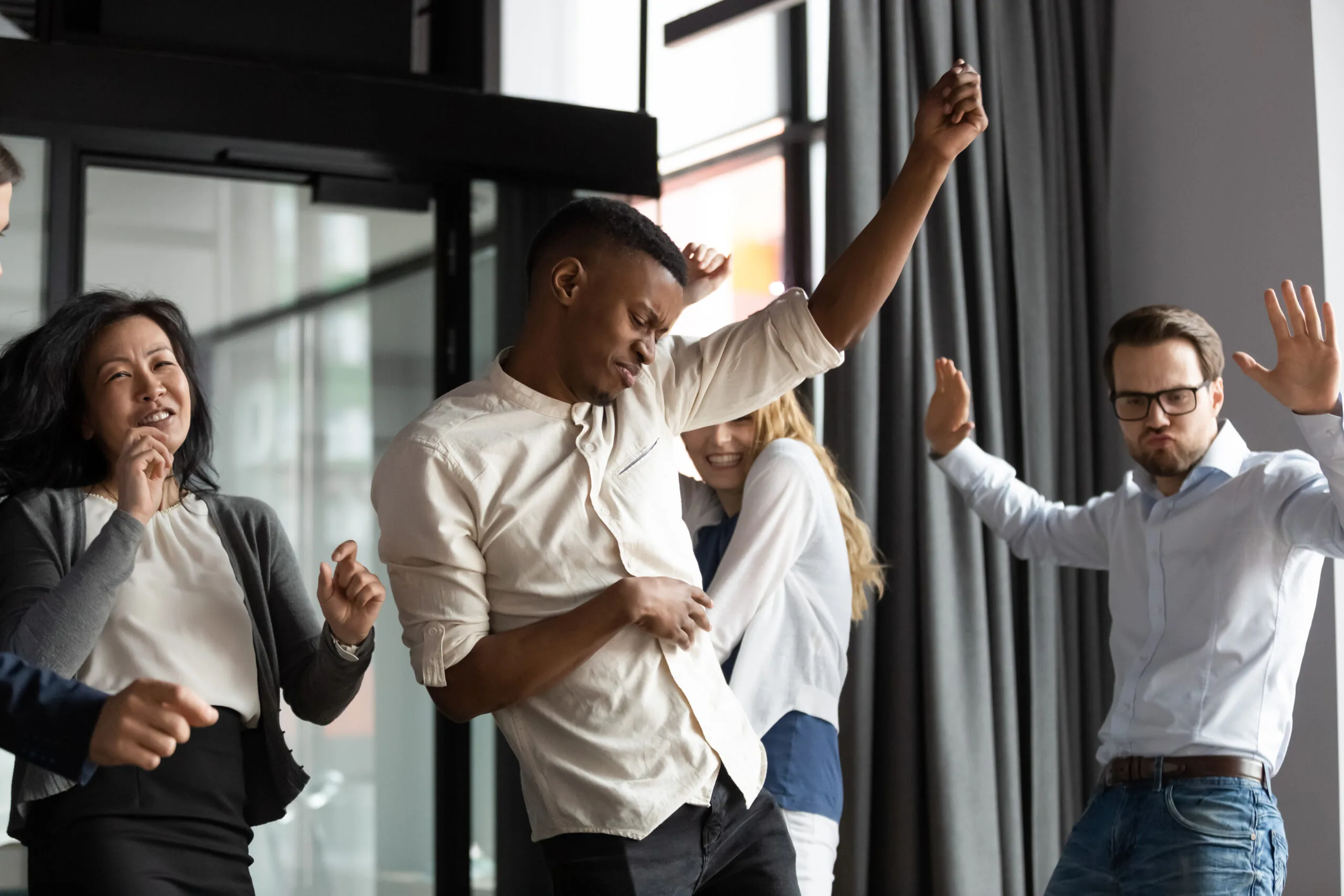 Group of colleagues dancing and celebrating with raised arms in a bright office near large windows