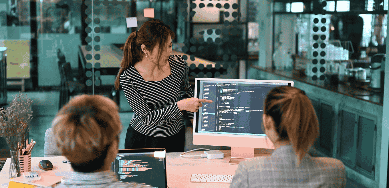 Woman in a striped top pointing at code on a monitor while two colleagues follow from their desks