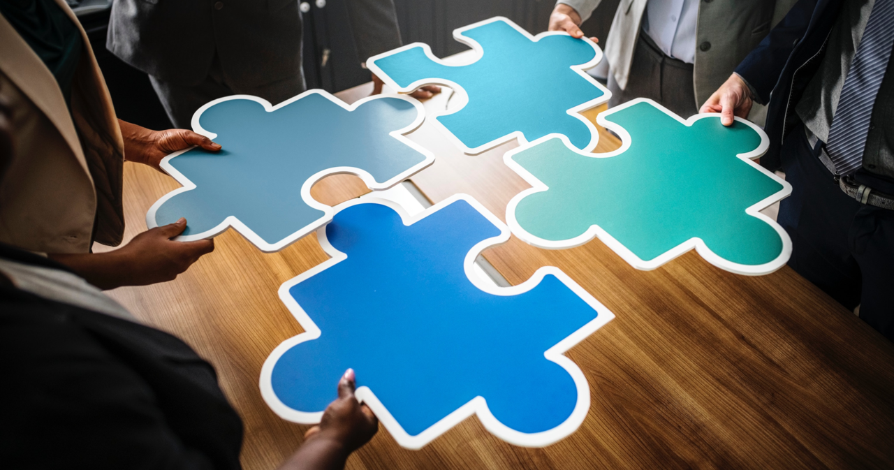 Four people fitting together large blue and green jigsaw puzzle pieces on a wooden table