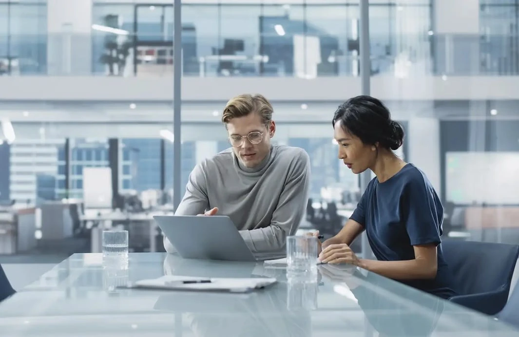 Man with glasses and a woman reviewing a laptop together at a glass table in a modern office