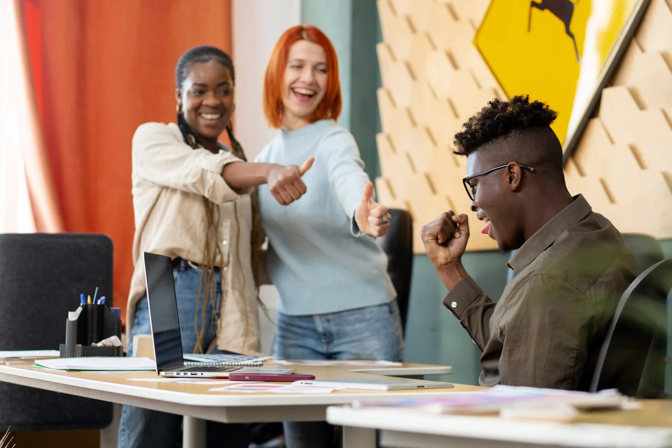 Two smiling women giving a thumbs up to a man celebrating in front of a laptop in an office