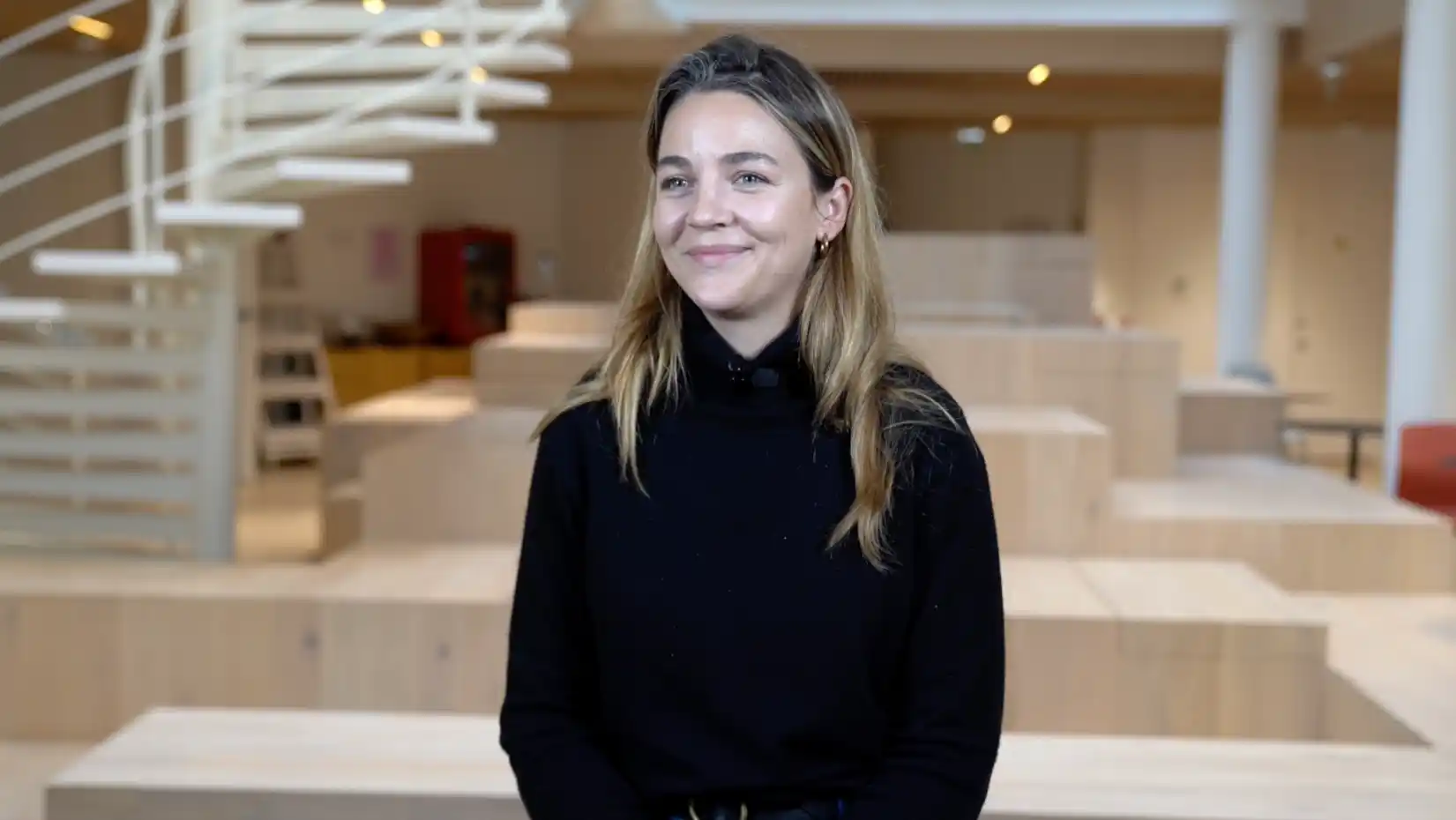 Smiling woman in a black sweater interviewed in a bright office with wooden bleacher seating and a staircase