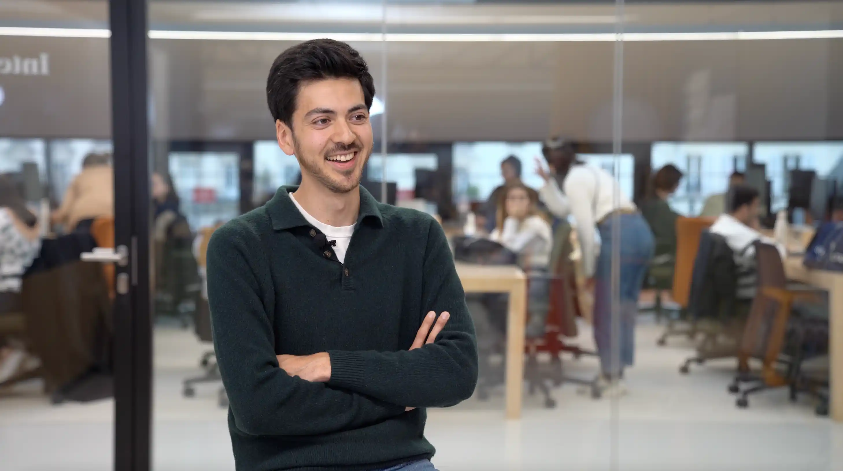 Smiling young man in a dark green sweater with arms crossed in front of a busy open-plan office