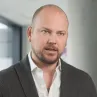 A bald man with a trimmed beard wearing a black blazer over a white shirt, standing indoors near a window with light gray walls in the background.