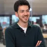 A smiling young man with short dark hair and a trimmed beard wearing a dark green sweater over a white collared shirt, standing with arms crossed in an office environment.
