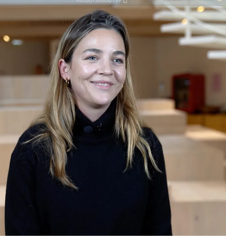 Smiling woman with light brown hair wearing a black turtleneck sweater in a softly lit indoor setting with blurred beige and wooden background elements.