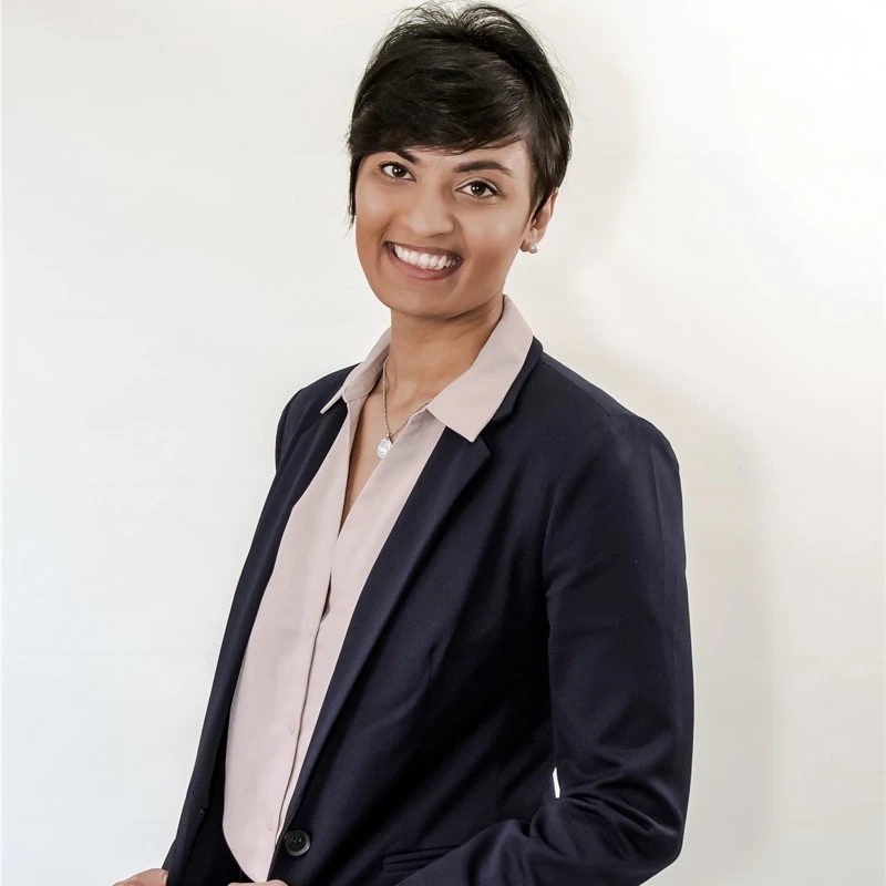 Smiling woman with short dark hair wearing a navy blazer over a light beige blouse, standing against a plain white background.