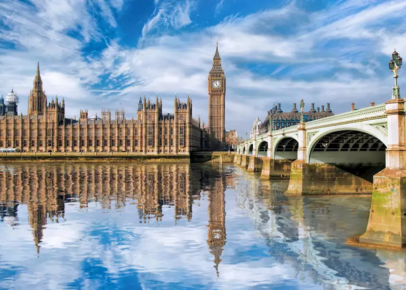 The Palace of Westminster with the clock tower known as Big Ben and Westminster Bridge reflected in the River Thames under a blue sky with wispy clouds.