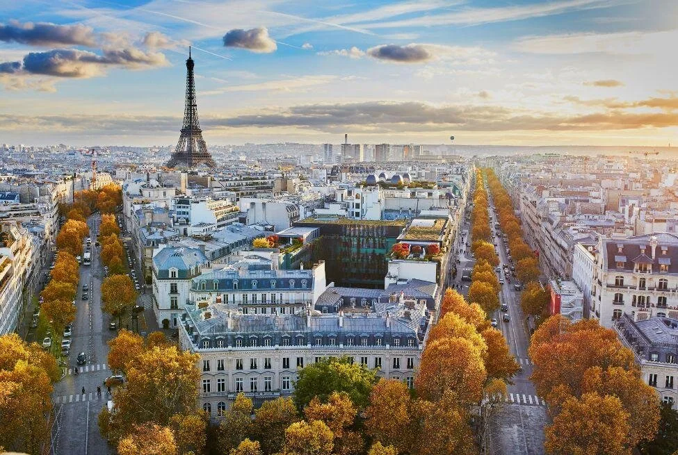 View of Paris at sunset with the Eiffel Tower prominently in the background and tree-lined avenues with autumn foliage in the foreground, surrounded by classic Parisian buildings.