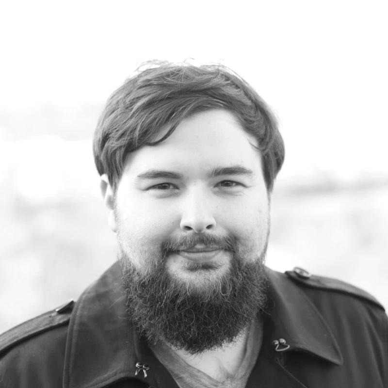 Black and white close-up portrait of a smiling man with a full beard and medium-length hair wearing a dark jacket.