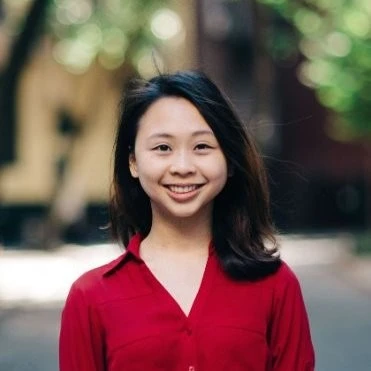 Smiling young woman with medium-length dark hair wearing a red blouse, standing outdoors with blurred urban background.