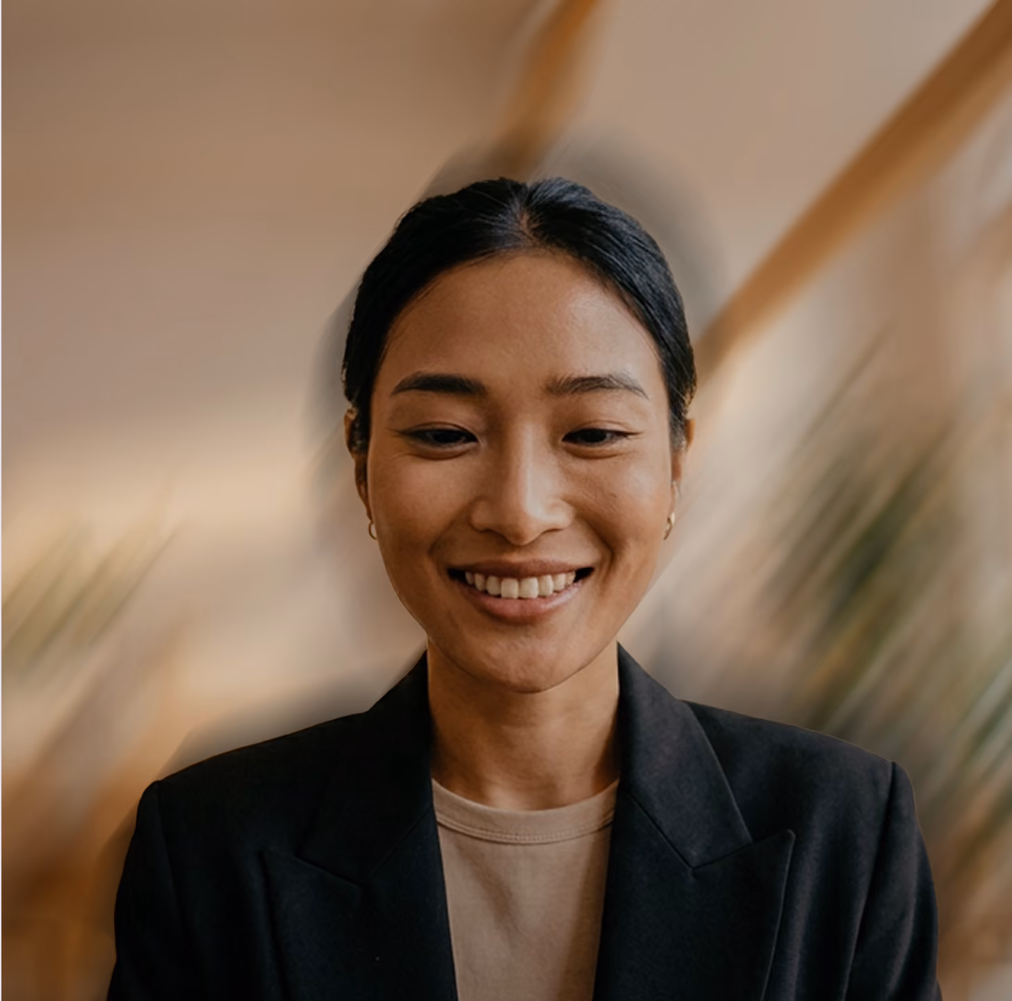 Smiling Asian woman with dark hair pulled back, wearing a black jacket and beige top, with a blurred warm-toned background.