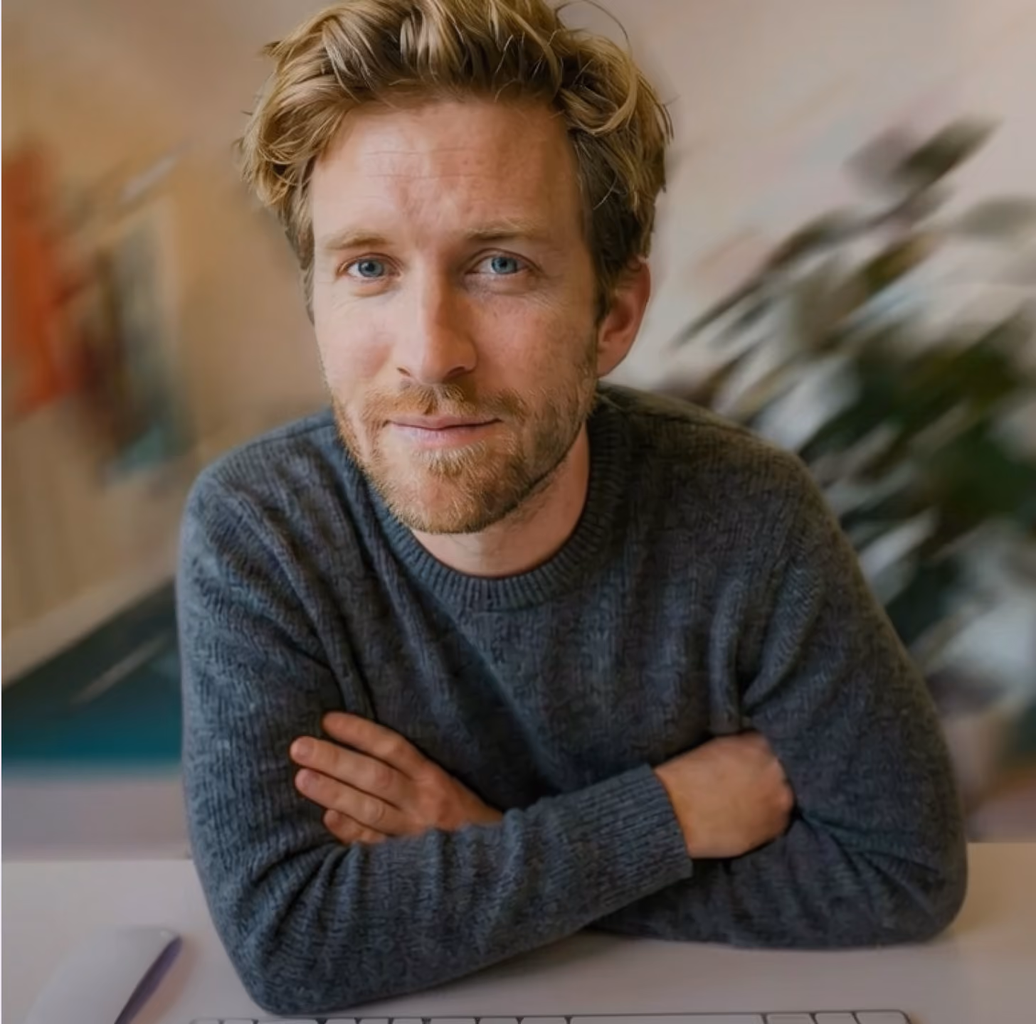 Man with blond hair and blue eyes wearing a gray sweater, sitting at a desk with arms crossed, looking at the camera in an indoor setting with blurred background.