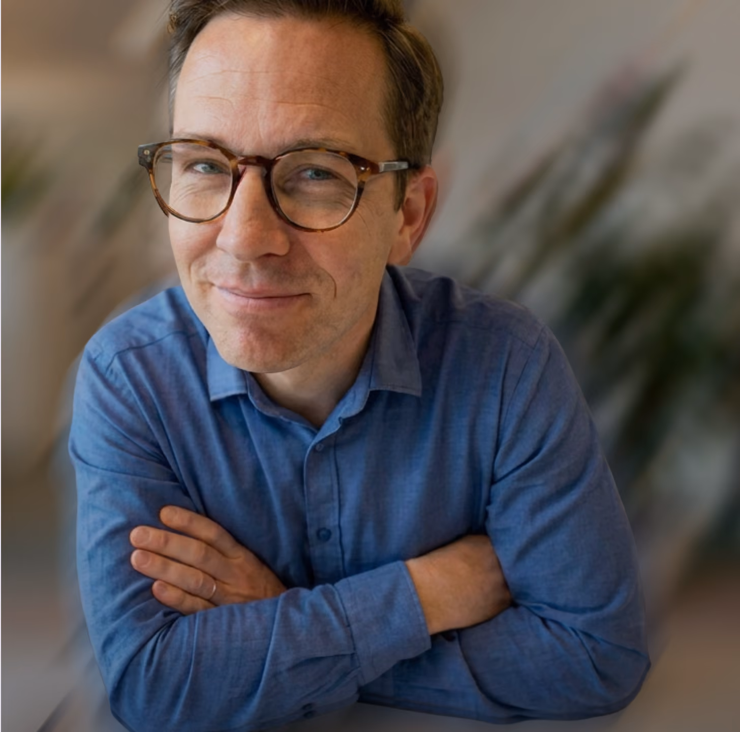Smiling man with short brown hair, wearing tortoiseshell glasses and a blue button-up shirt, sitting with arms crossed in a blurred indoor setting.