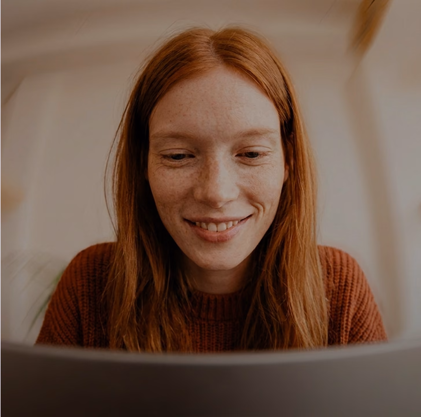 Red-haired woman wearing a brown sweater smiling and looking at a laptop screen indoors.