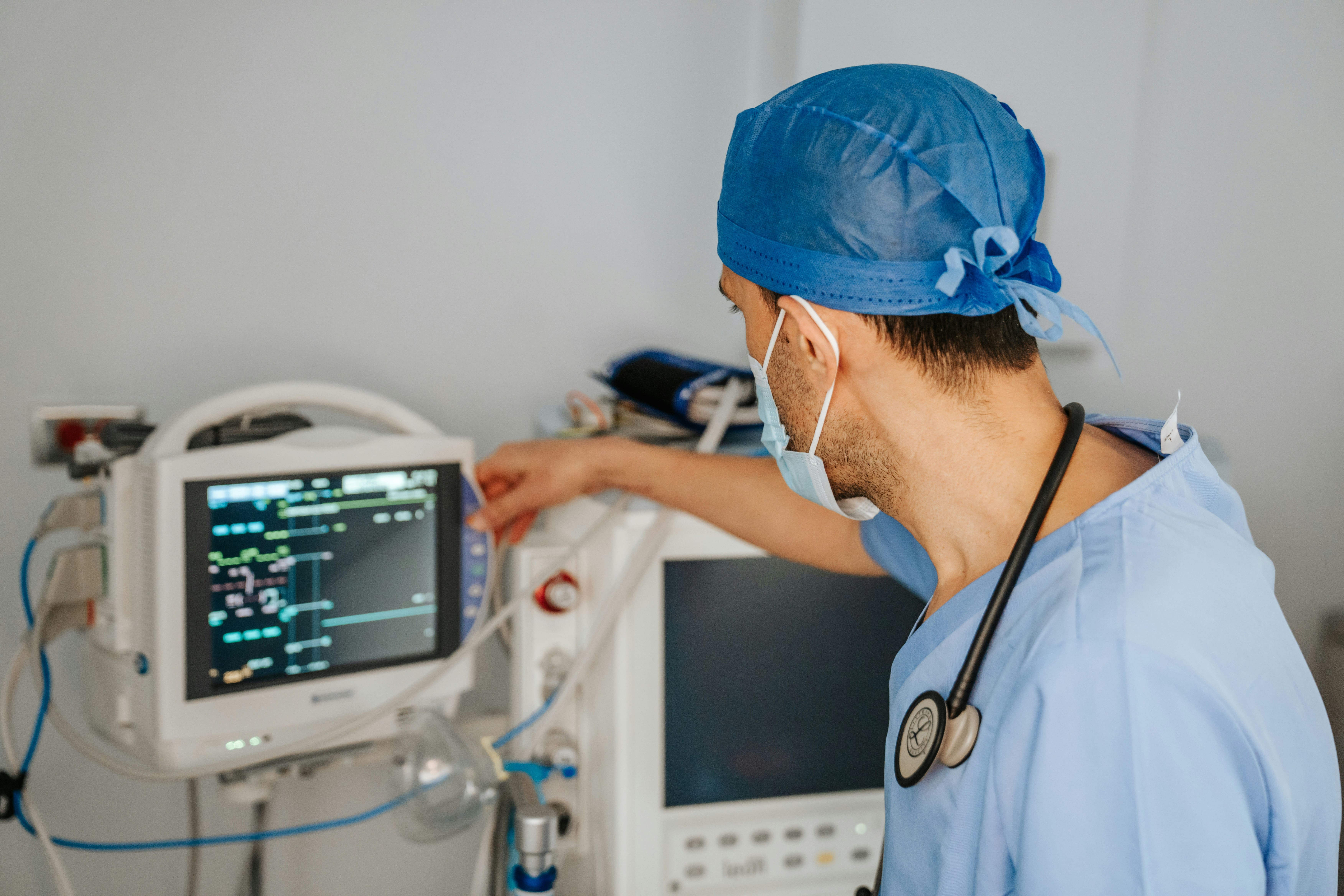Healthcare professional in surgical scrubs, mask, and cap adjusting settings on a medical monitor with a stethoscope around their neck in a clinical environment.