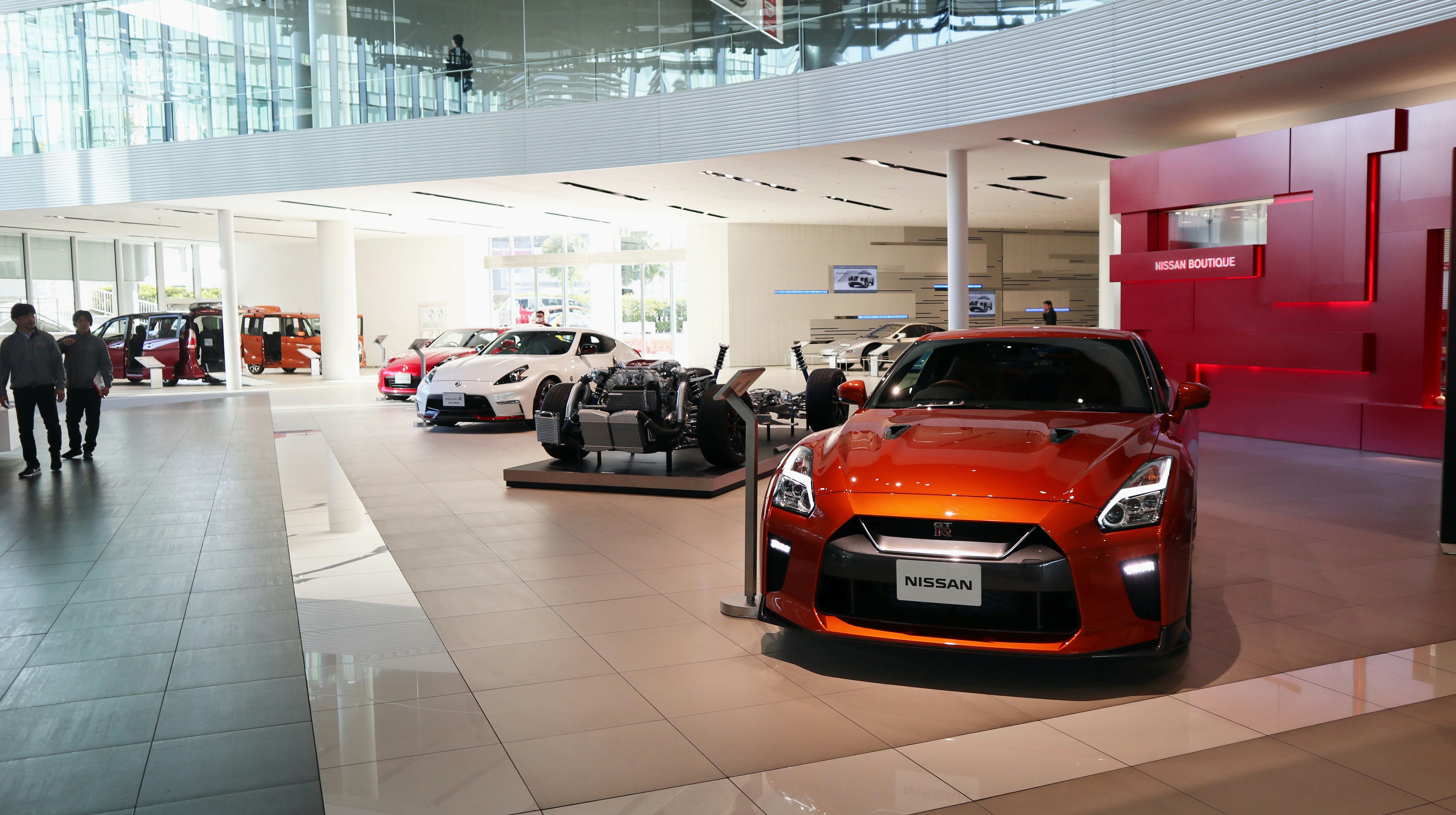 Modern Nissan car showroom with a bright orange Nissan GT-R prominently displayed on the right, alongside other Nissan sports cars and a car chassis model in the background, featuring sleek glass architecture and a red wall labeled Nissan Boutique.