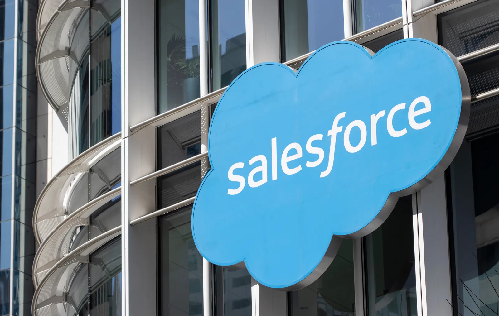 Blue cloud-shaped sign with the white text 'salesforce' mounted on the exterior of a modern glass and metal building.
