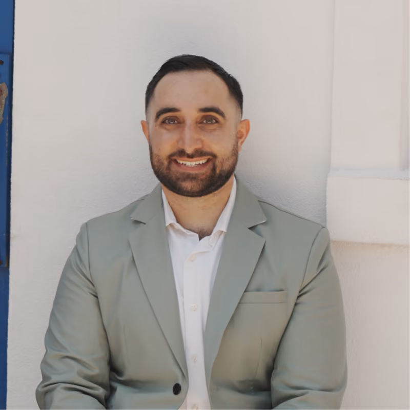 Smiling man with short dark hair and beard wearing a light green blazer and white shirt, sitting against a white wall with a blue edge on the left.