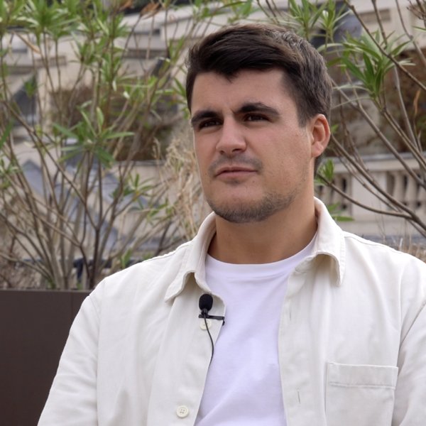 Young man with short dark hair wearing a white shirt and jacket seated outdoors with greenery and blurred buildings in the background.