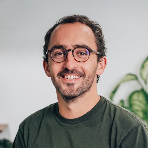 Smiling man with short curly hair, beard, and glasses wearing a dark green shirt, with a blurred plant in the background.