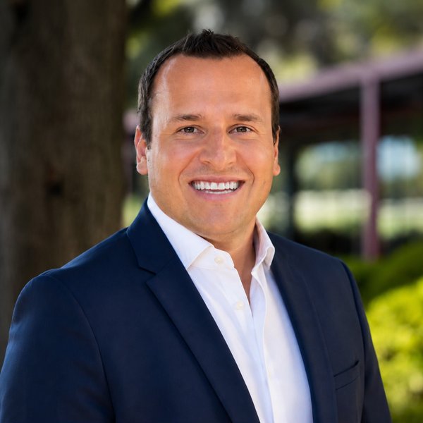 Smiling man wearing a dark blue suit jacket and white dress shirt standing outdoors with trees and greenery in the blurred background.
