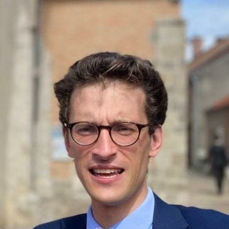 A young man with dark curly hair and glasses wearing a blue suit jacket and light blue shirt, standing outdoors with buildings and a narrow street in the background.
