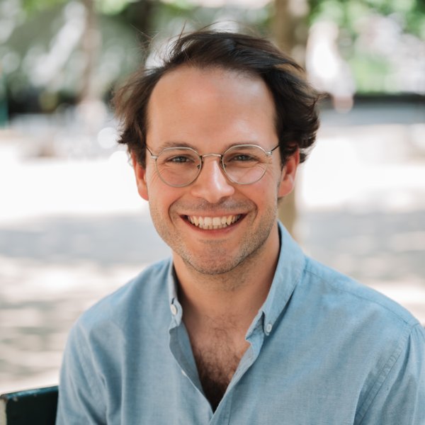 Smiling man with glasses and thinning hair wearing a light blue button-up shirt outdoors in soft daylight with a blurred background.