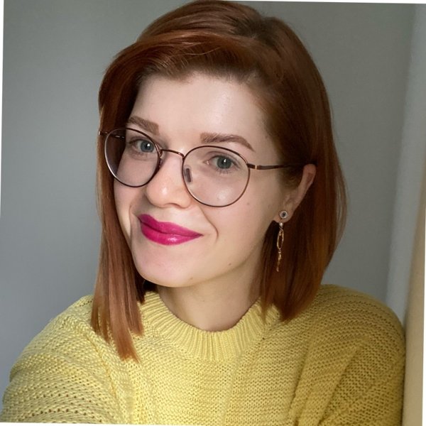 Woman with straight red hair wearing round eyeglasses, bright pink lipstick, a yellow knitted sweater, and a dangling earring, smiling slightly against a plain background.