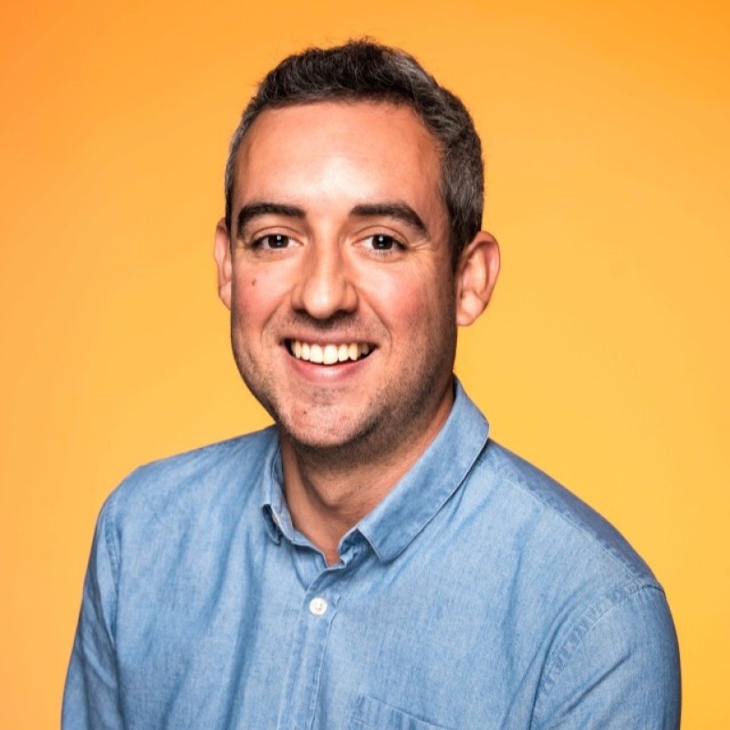 Smiling man with short dark hair and light stubble wearing a blue button-up shirt against an orange background.