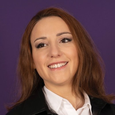 Smiling woman with long brown hair wearing a white shirt and black jacket against a purple background.