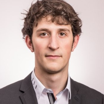 Portrait of a young man with curly brown hair wearing a dark blazer and a light-colored collared shirt against a plain background.