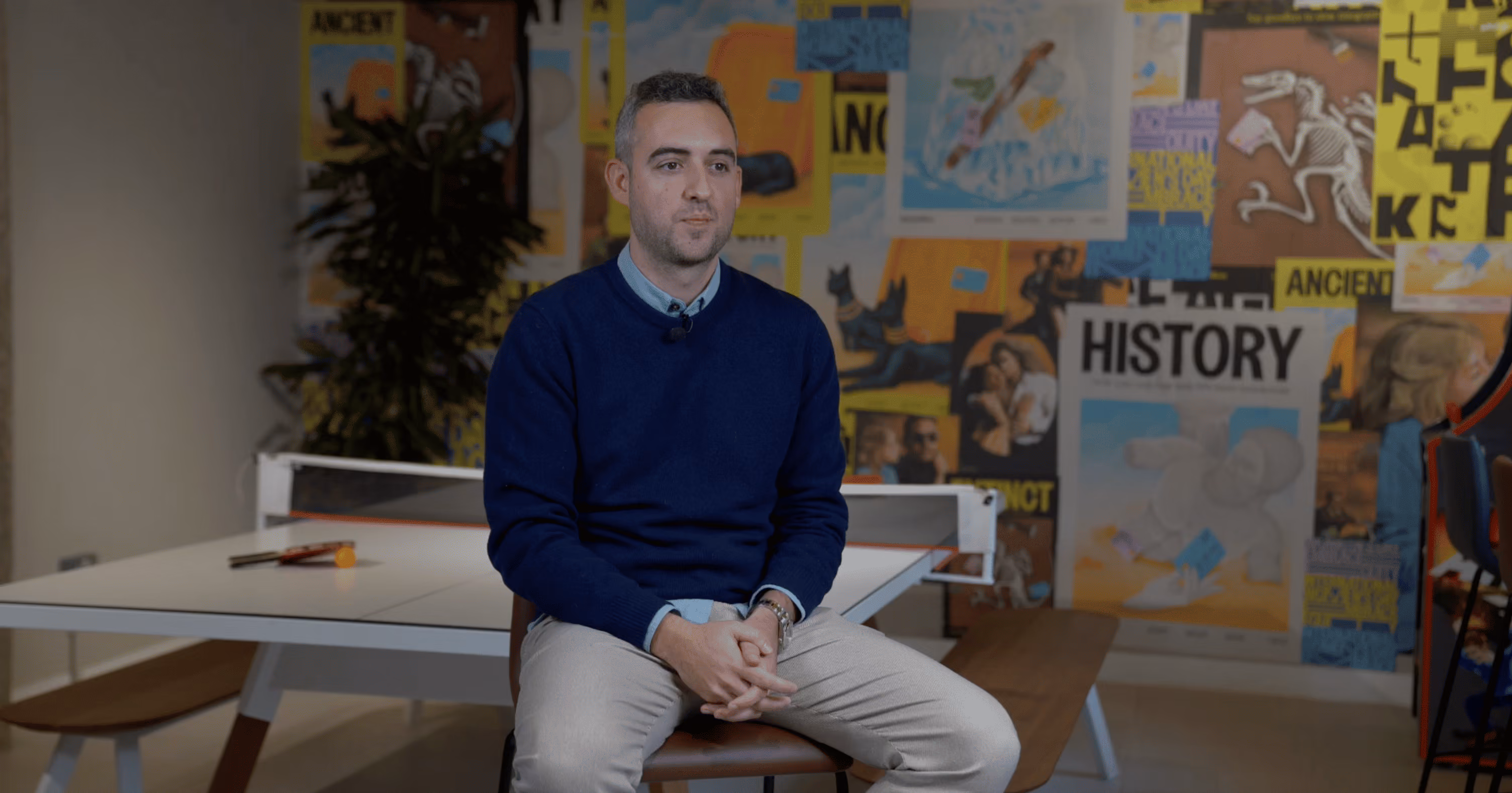 Man in a blue sweater sitting on a chair in front of a ping pong table with a colorful collage of posters and artwork on the wall behind him.