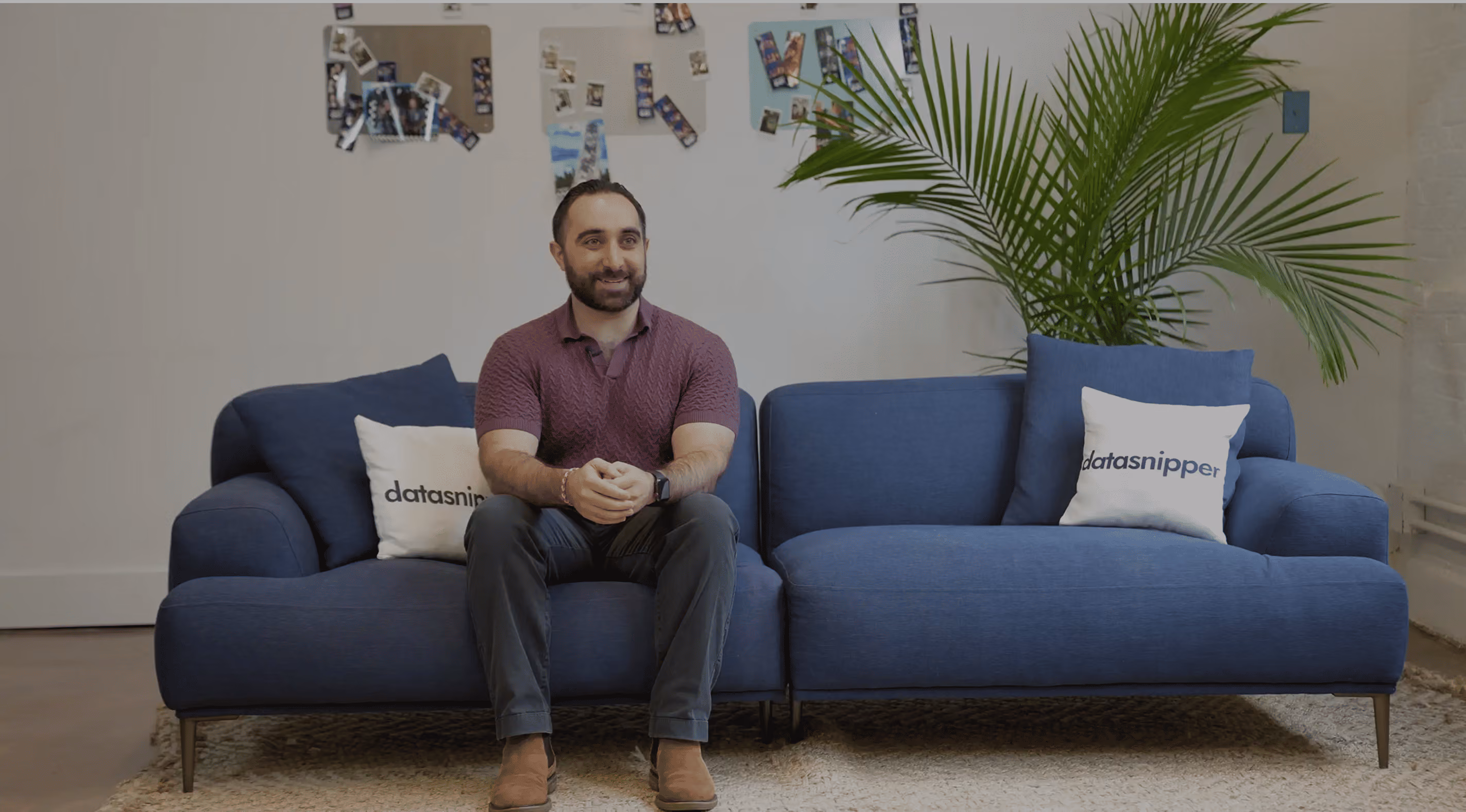 A man with a beard wearing a purple shirt and dark pants sitting on a blue sectional sofa with two white pillows labeled 'datasnipper' in a room with a large green plant and a wall decorated with photos.