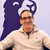 Smiling man with glasses and short dark hair wearing a light beige quarter-zip sweater, sitting in front of a purple backdrop with a lion head logo.