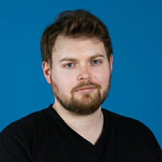 Portrait of a young man with light skin, brown hair, and beard wearing a black V-neck shirt against a blue background.
