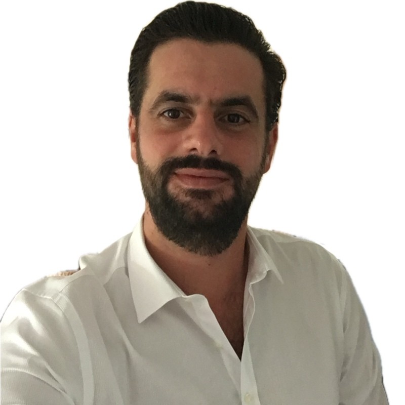 Man with dark hair and beard wearing a white button-up shirt, smiling slightly against a white background.