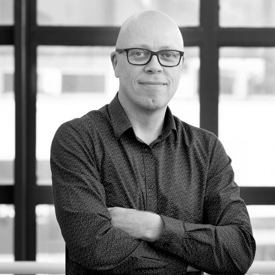 Black and white portrait of a bald man with glasses wearing a patterned button-up shirt, standing with arms crossed in front of a window.