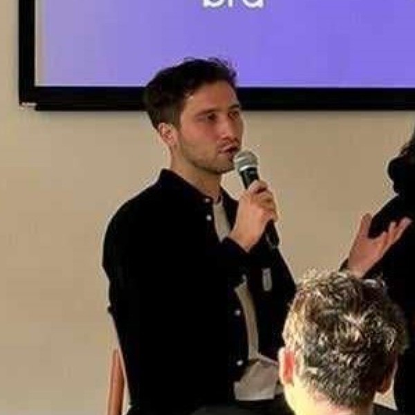 Man with dark hair speaking into a microphone during an indoor event, with an audience member visible from behind in the foreground.