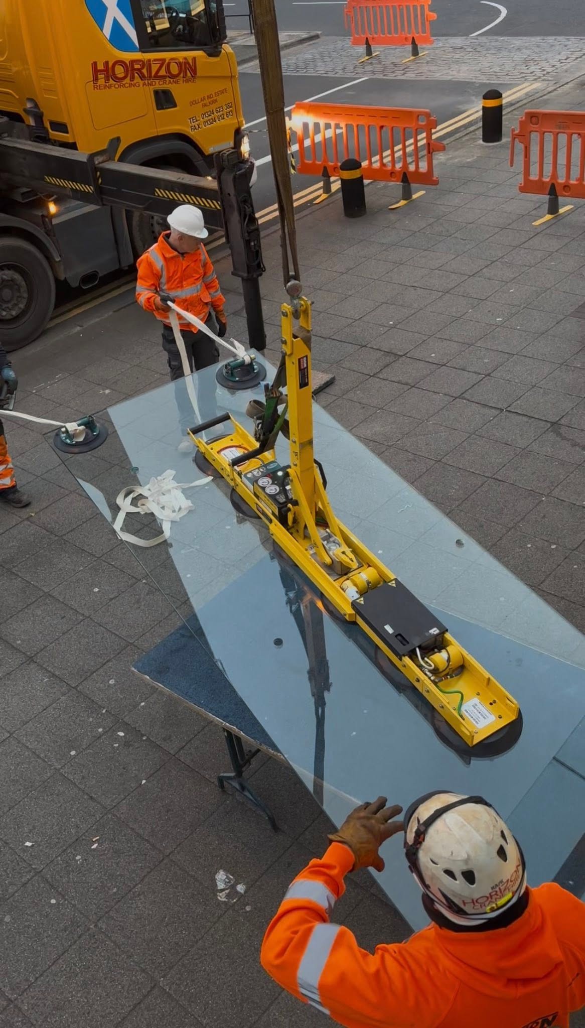 Large glass panel lifted by vacuum crane with workers in orange hi-vis on site
