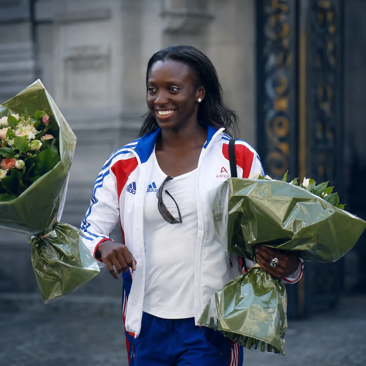 Smiling athlete in white Adidas jacket holding two bouquets of flowers outdoors.
