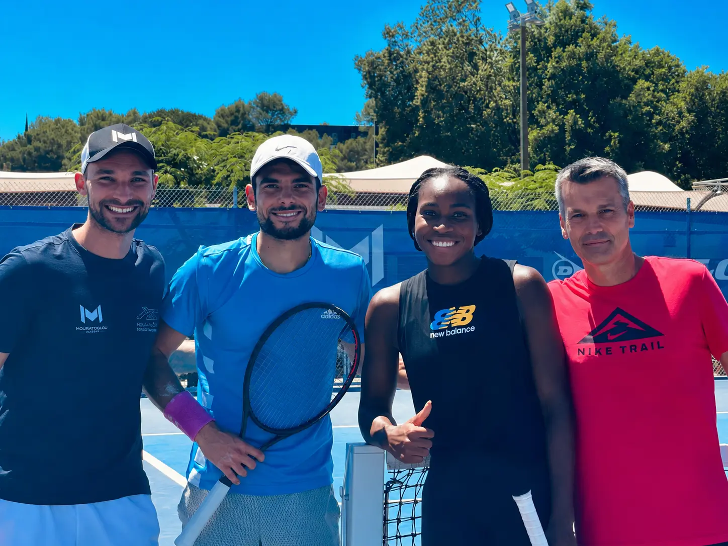 Four tennis players posing on blue court, one holding racket, one giving thumbs up, all smiling.