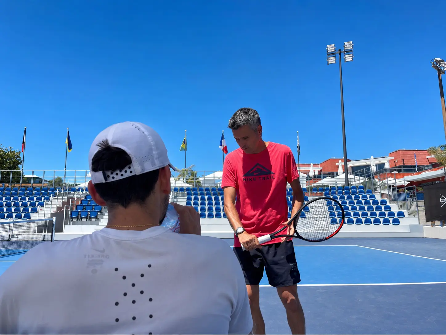 Tennis coach instructs player holding racquet on a blue court with empty stadium seats.