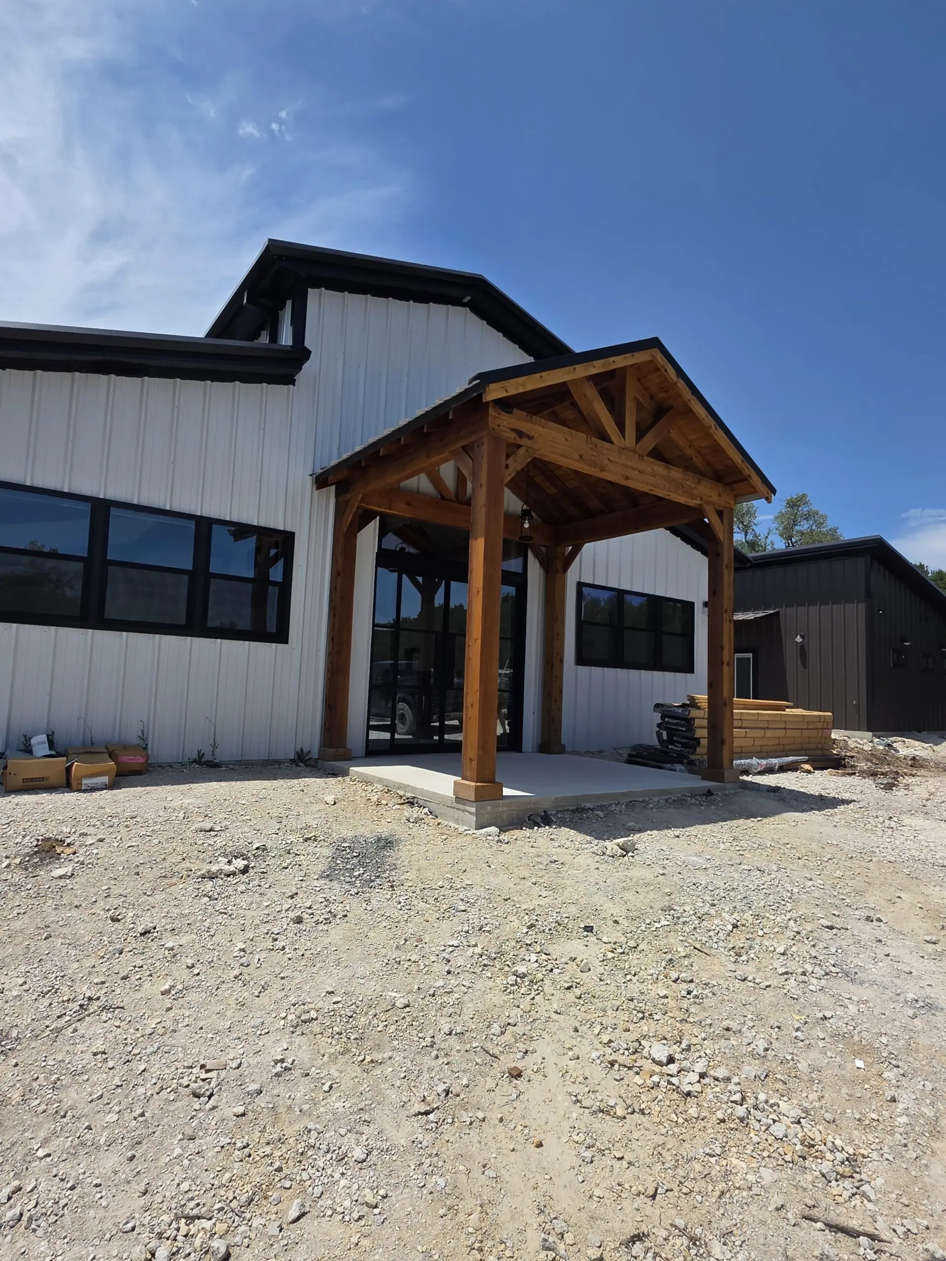 White metal barndominium with black trim and a cedar timber entry porch, nearing completion in the Texas Hill Country