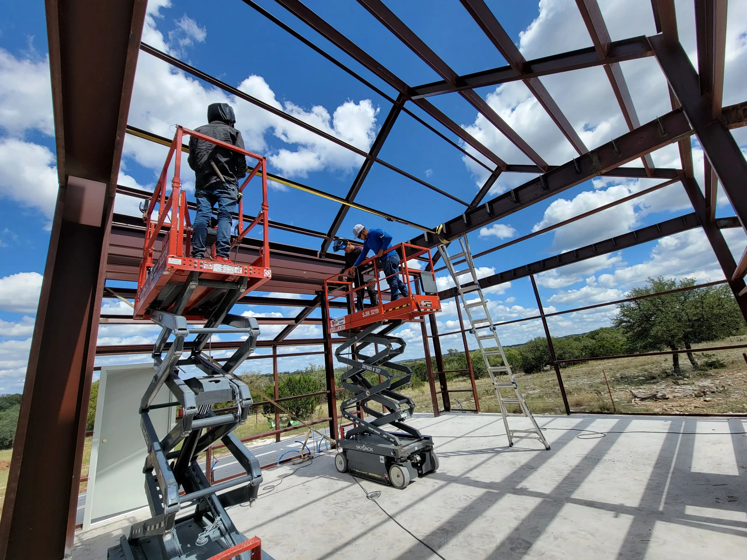 Two crew members on scissor lifts assembling the steel frame of a building on a Hill Country slab