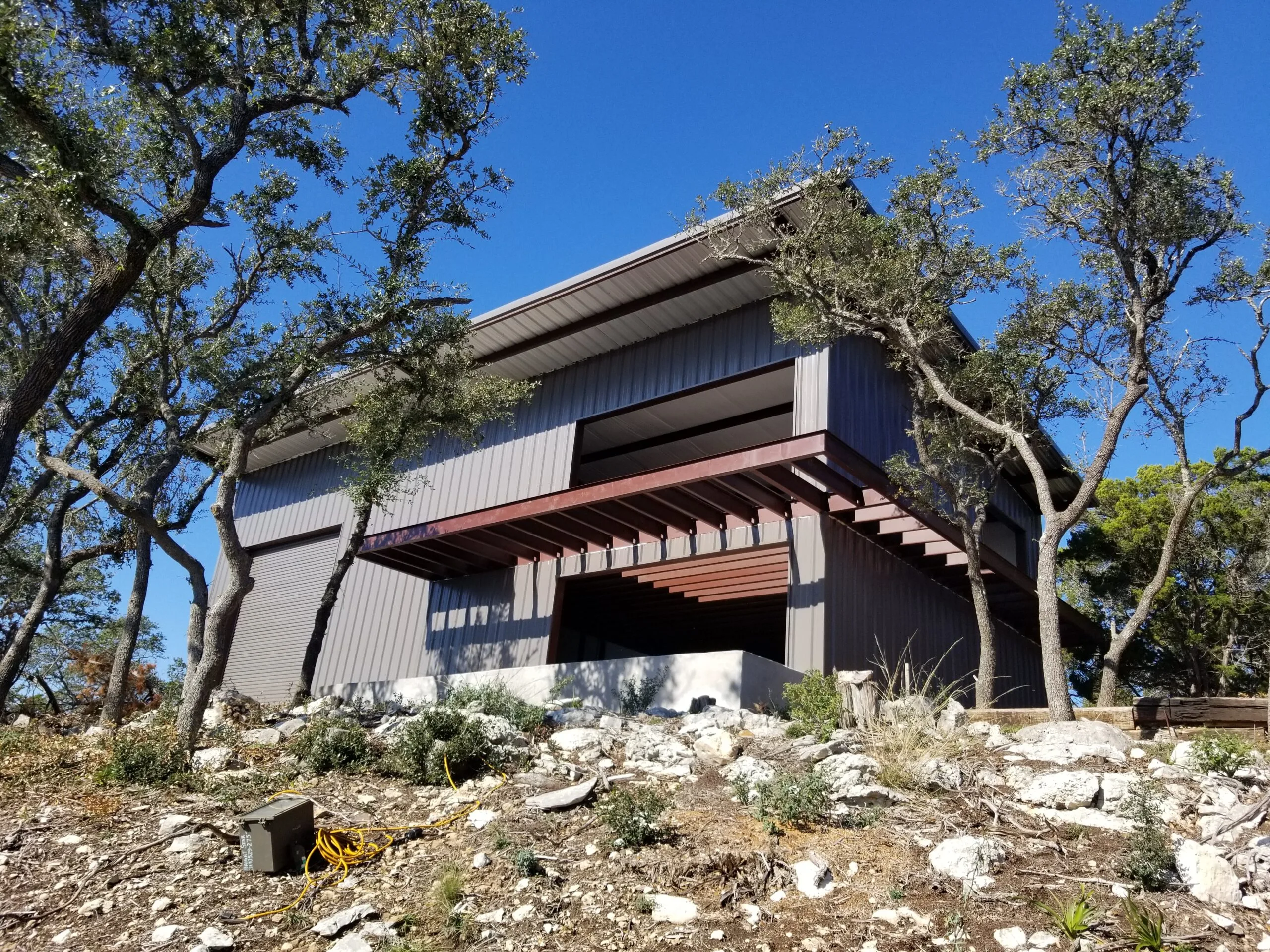 Grey steel barndominium with a cantilevered covered porch and exposed steel beams on a rocky oak-covered Hill Country lot