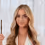 Smiling woman with long blonde hair holding silver tongs against white background.